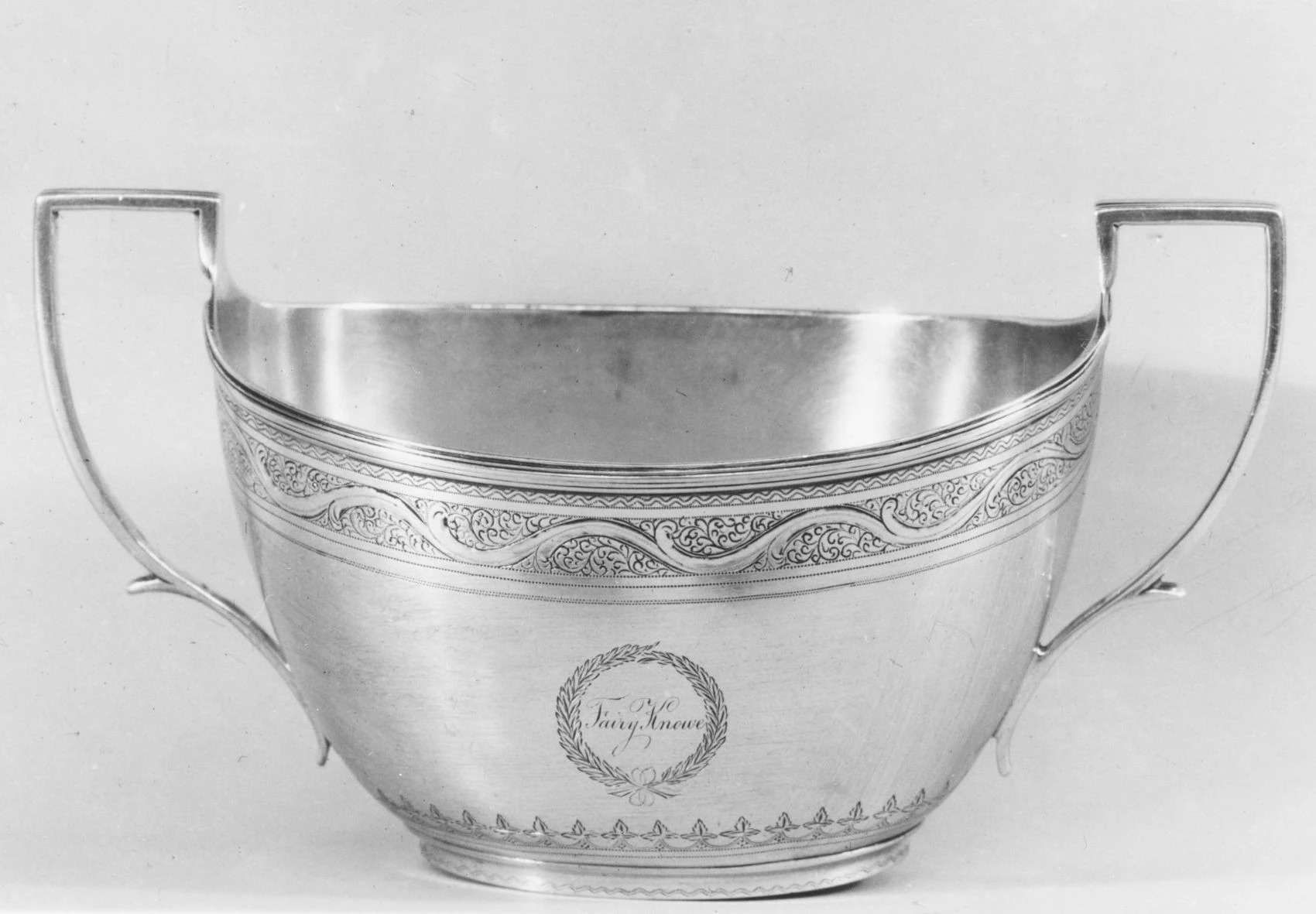 A two-handled sterling silver sugar bowl with a chased scroll-and-leaf border below the rim and an engraved wreath cartouche reading "Fairy Knowe" on the front