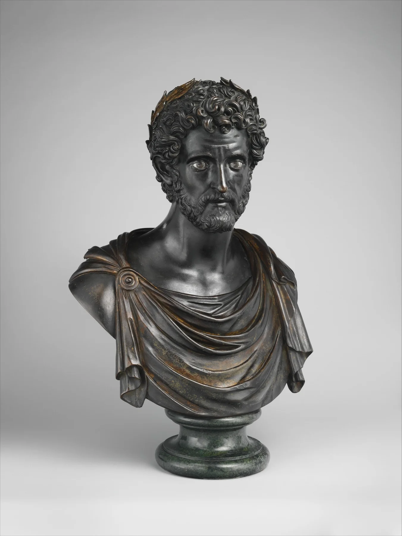 A bronze bust of the Roman emperor Antoninus Pius, dated 1519 to 1524, showing a bearded draped figure with visible warm patina and gilt highlighting on the drapery, mounted on a dark green stone socle