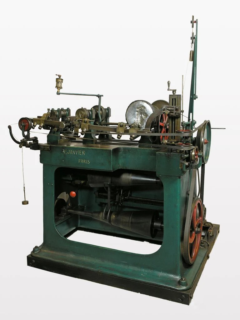A green-painted Janvier reducing lathe, a pantograph machine with a sculpted portrait model mounted on one side and cutting tools on the other, used to transfer a design onto a coin die at reduced scale