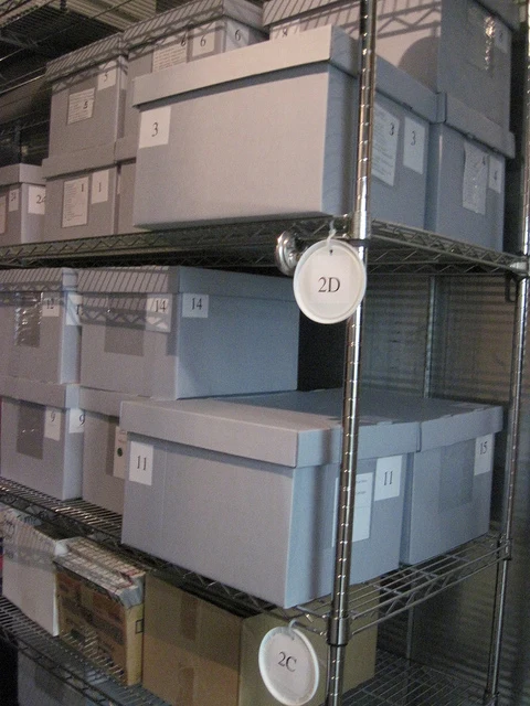Rows of numbered, labeled grey archival storage boxes stacked on metal wire shelving in a museum collections storage room