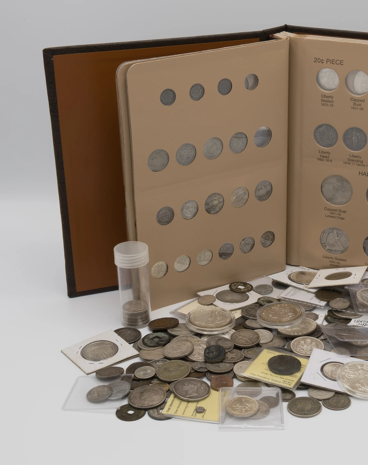 An open coin album with rows of coins in individually labeled slots, next to a plastic tube of coins and a large loose pile of assorted old coins in paper and plastic holders
