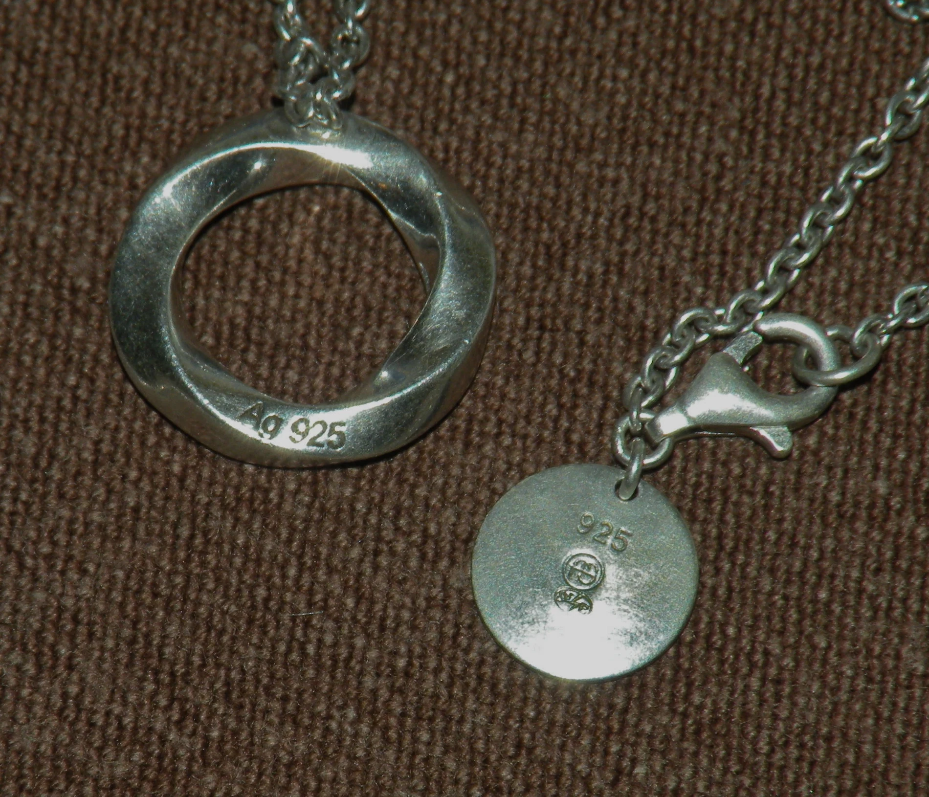 A smooth sterling silver ring pendant stamped "Ag 925" on its inner edge, next to a round silver tag stamped "925" beside a small maker's mark, both pieces resting on a woven fabric surface