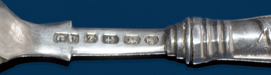 Close view of the shank of a British silver spoon struck with five hallmark punches in a row: a maker's mark reading "GU," a date letter, and three further assay symbols
