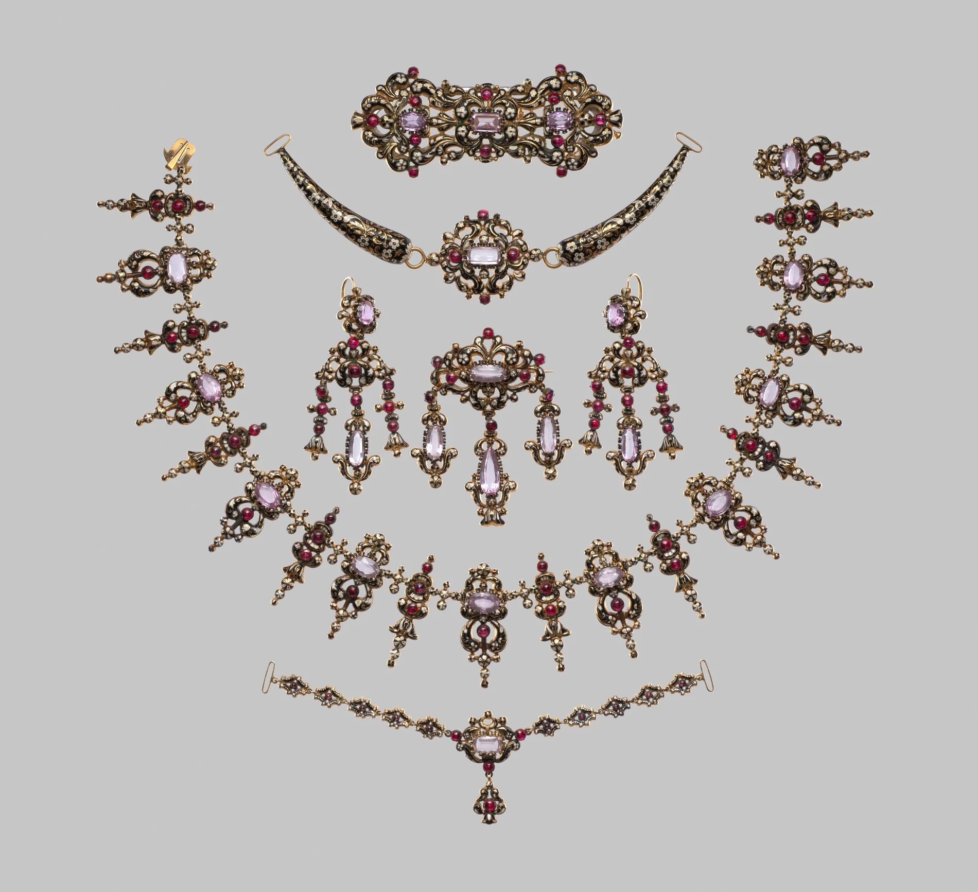 A complete Victorian parure laid out on a neutral grey ground: a fringed necklace, a matching bracelet, a large brooch, a pair of chandelier earrings, a hair ornament and a decorative comb, all in gold with black enamel and pale pink glass-paste stones
