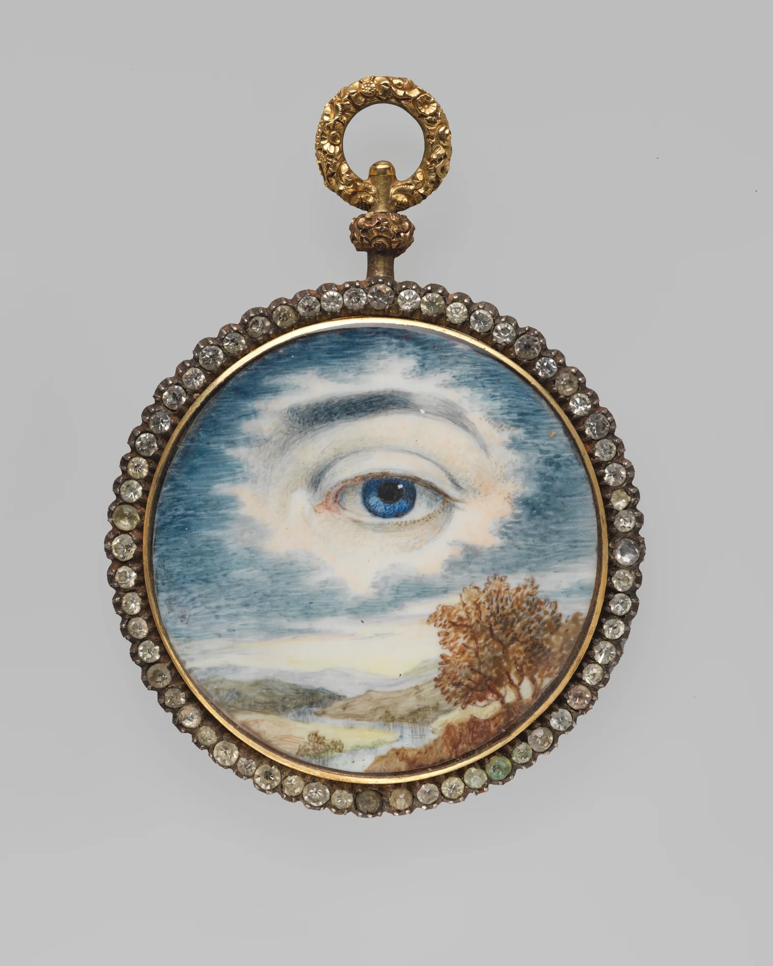 A round gold locket bordered with clear stones, set with a watercolor miniature of a single painted eye beneath a cloudy sky and a landscape with a tree, a lover's eye keepsake from about 1840