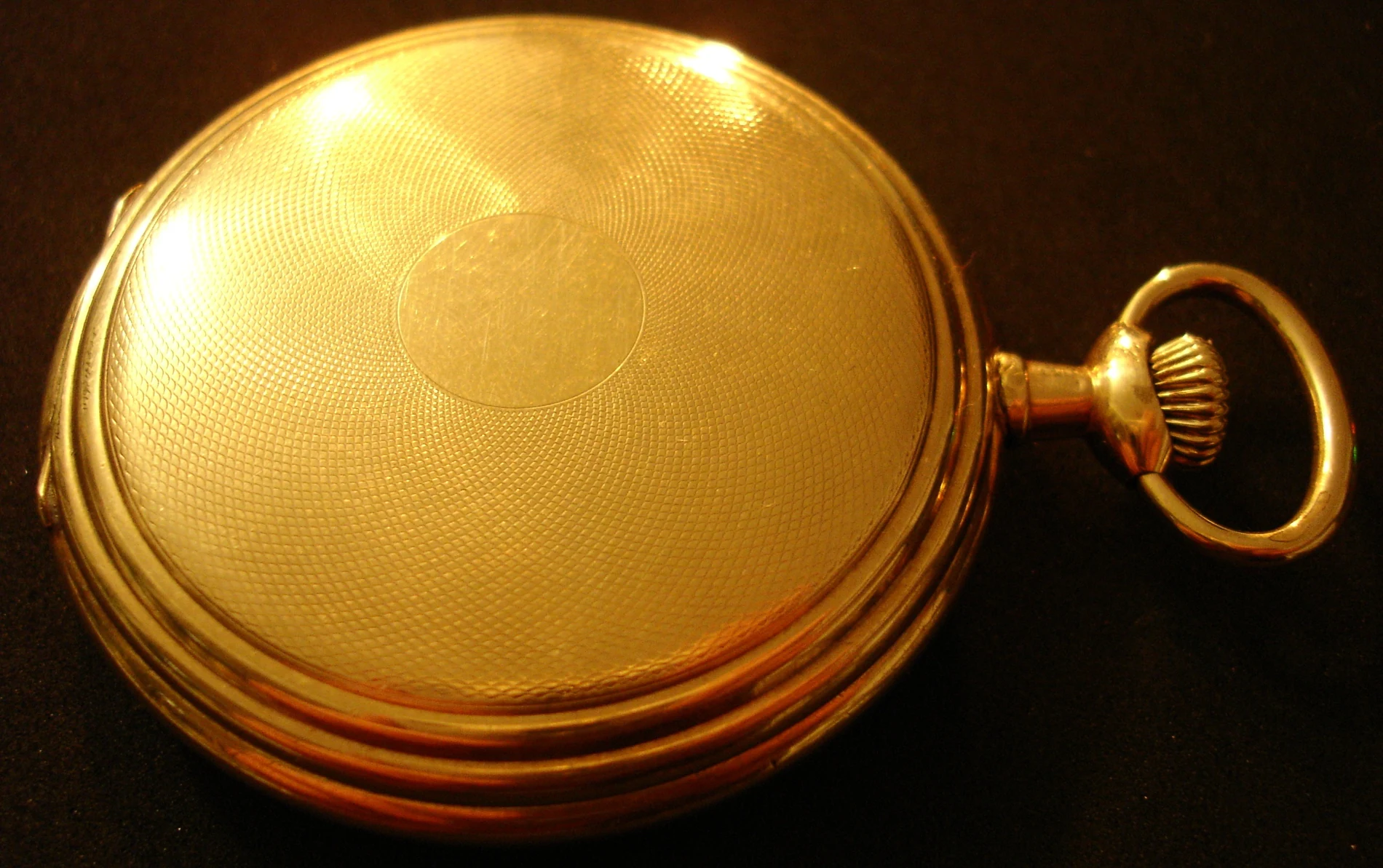 The closed gold case back of a 1911 Elgin pocket watch, covered edge to edge in a fine radiating engine-turned guilloché pattern around a blank oval cartouche, with the winding crown and bow visible at the right