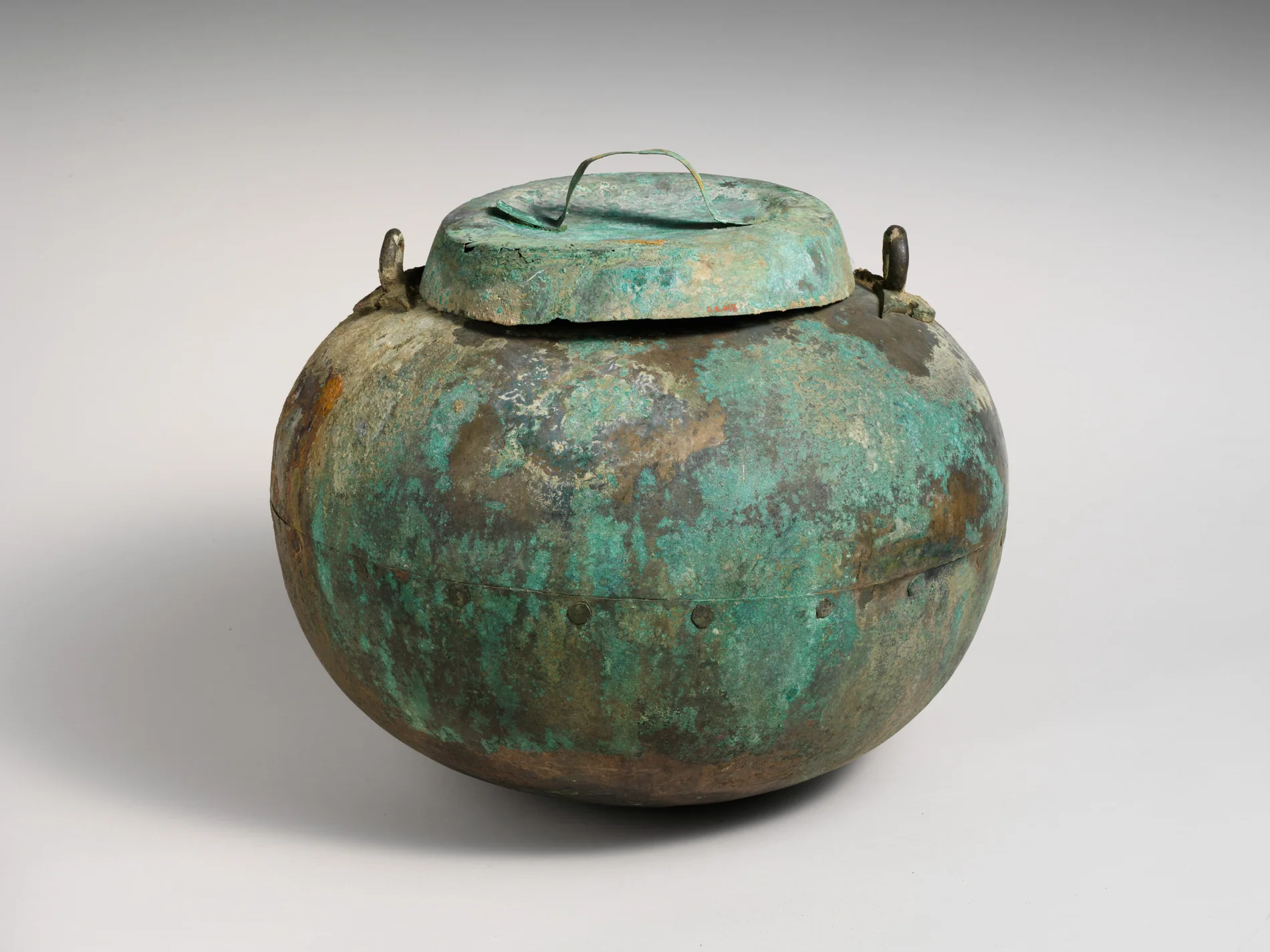 A round-bodied Etruscan bronze cauldron with a domed lid and looped handles, its entire surface covered in mottled green and brown corrosion built up over roughly twenty-five centuries