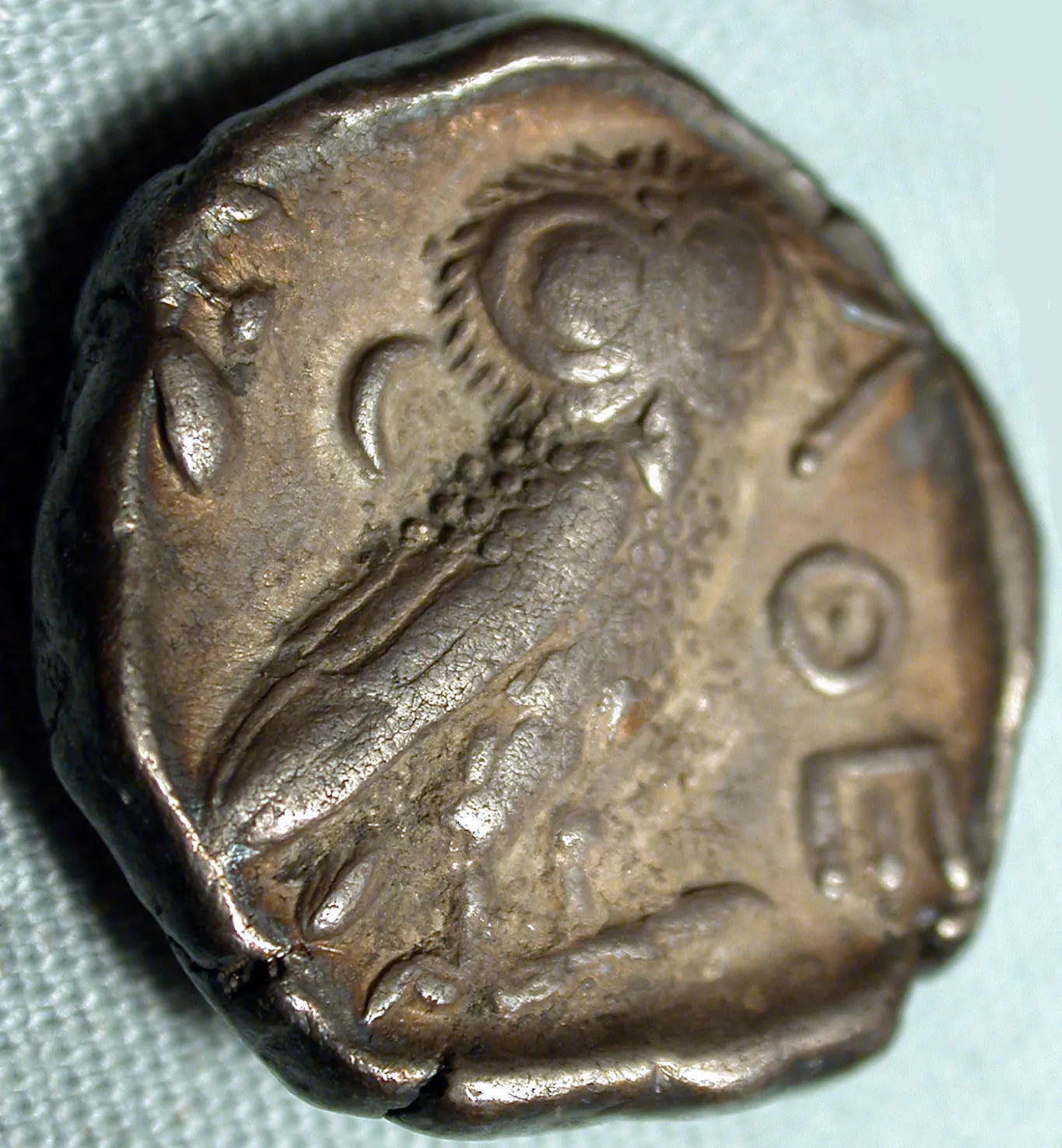 The reverse of an ancient Athenian silver coin, worn and toned, showing an owl in relief with an olive sprig and crescent moon above its head and traces of an inscription to the right