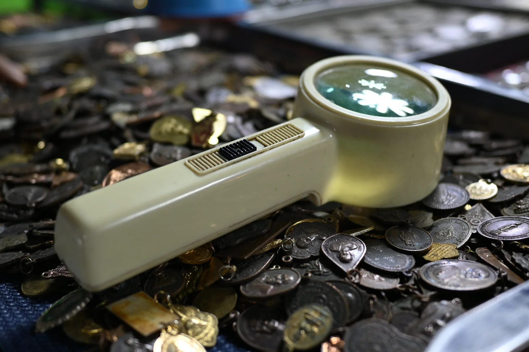 A handheld illuminated magnifying loupe resting on a scattered tray of old coins, medals and small metal tokens during close examination
